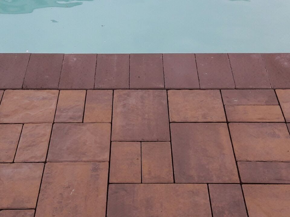 A close-up of brown rectangular patio pavers laid in a multi-size pattern along the edge of a swimming pool.