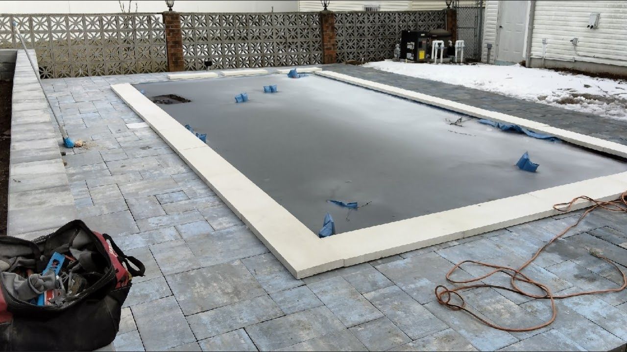 A backyard swimming pool covered for winter, surrounded by grey pavers, with a tool bag in the foreground.