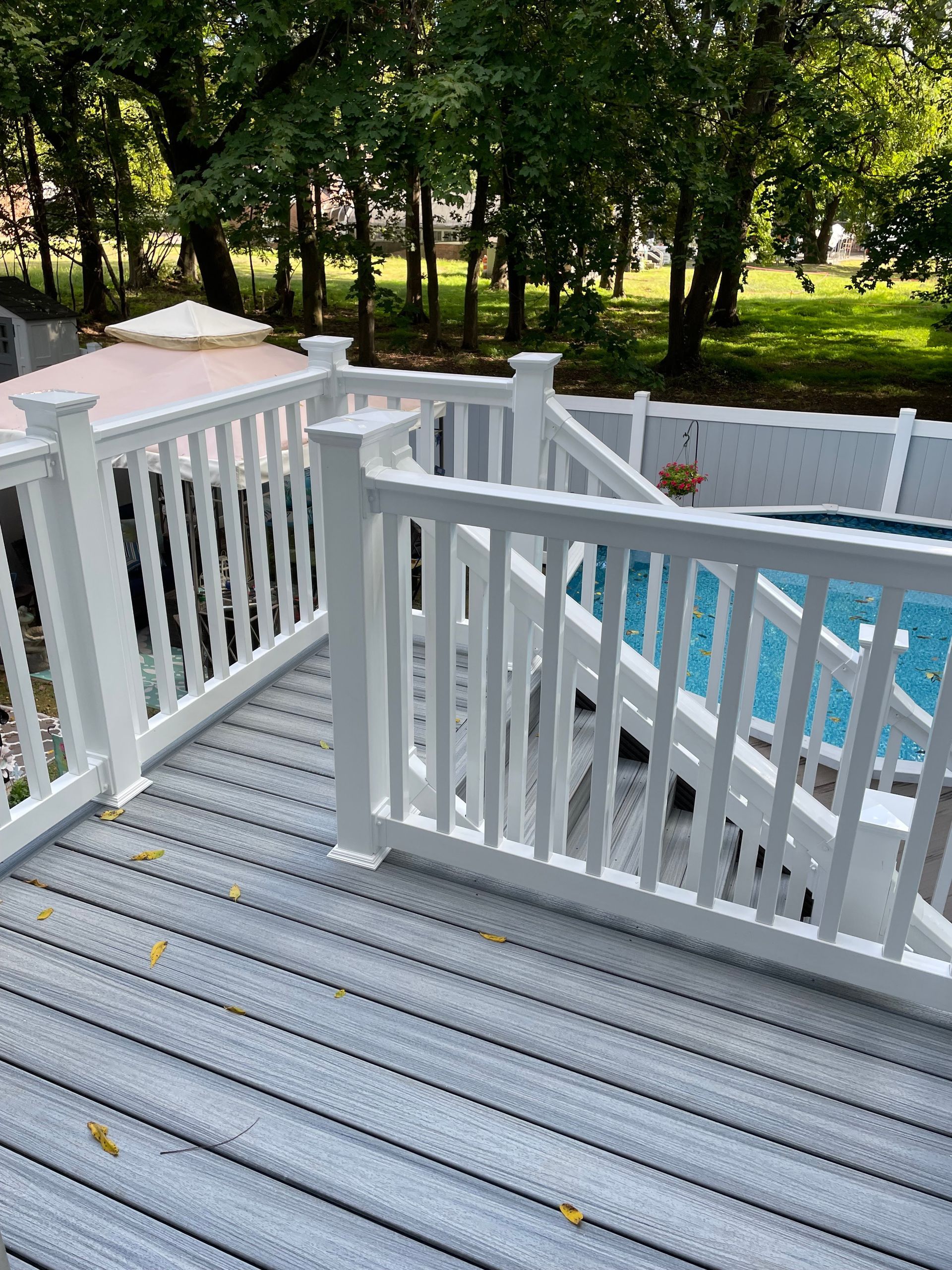 A gray composite deck with white railings overlooks a backyard swimming pool and trees on a sunny day.