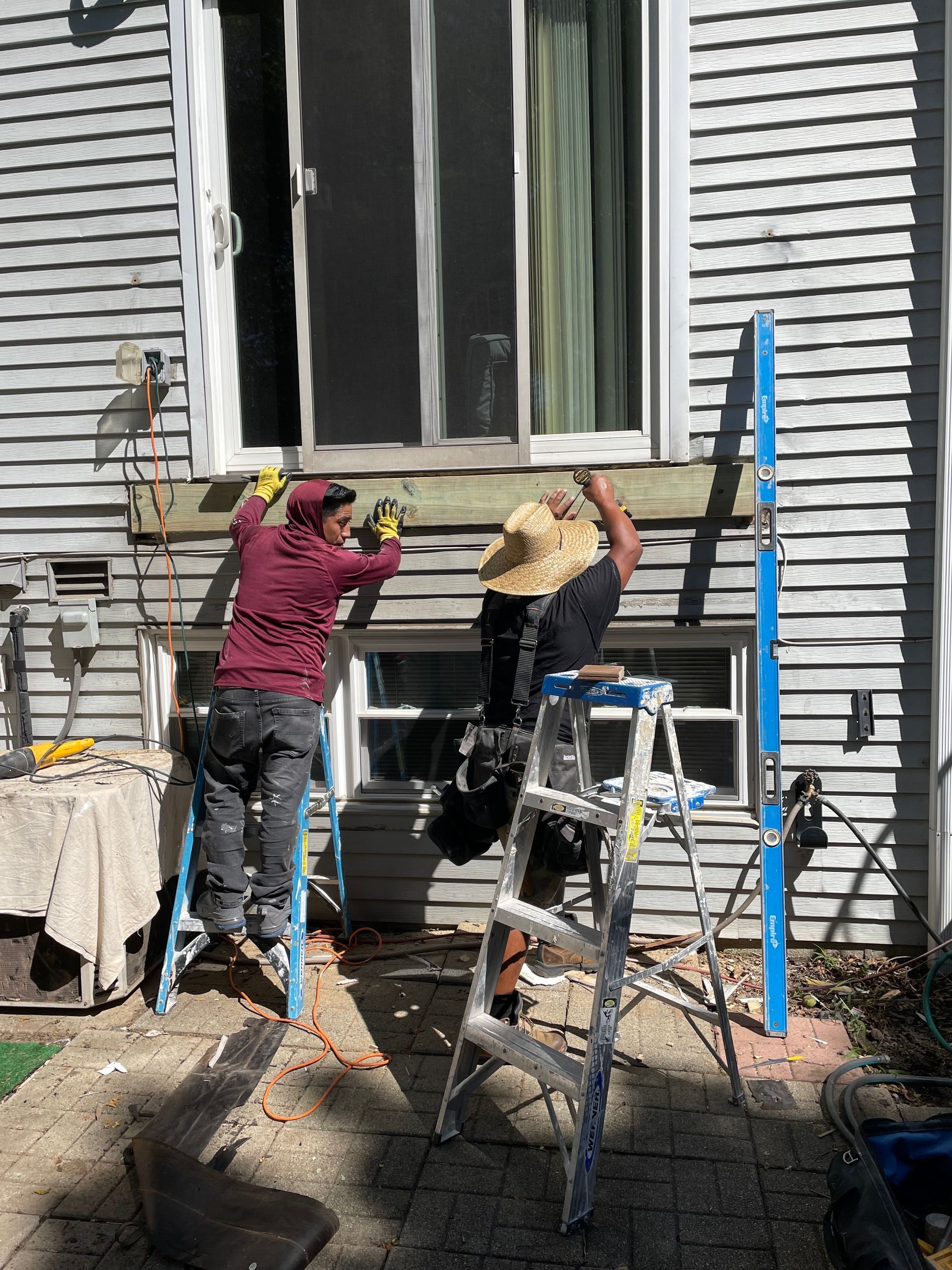 Two people on step ladders work on installing a new header board under a house window.