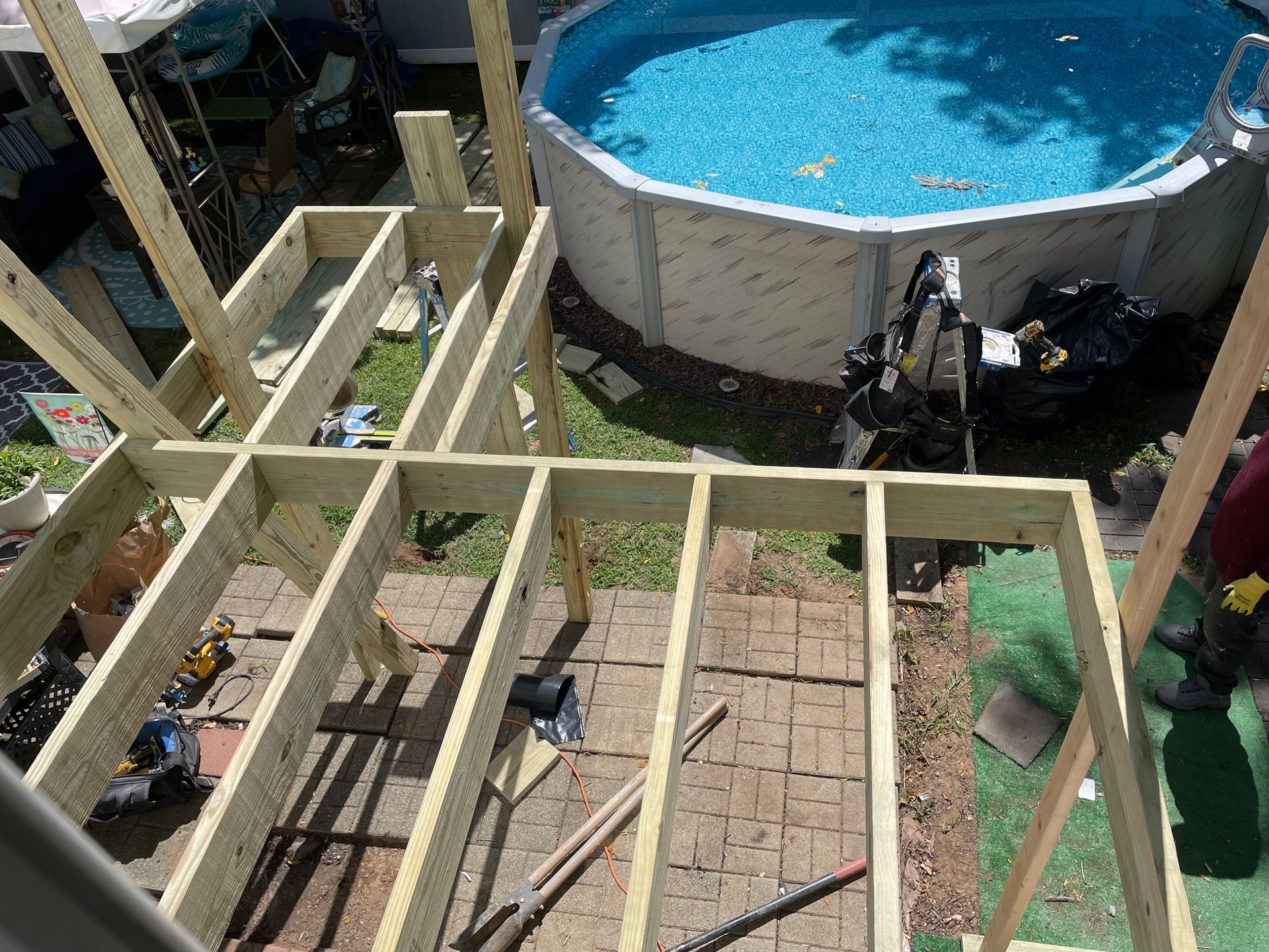 Wooden deck framing under construction above a brick patio, with a round above-ground pool in the background.