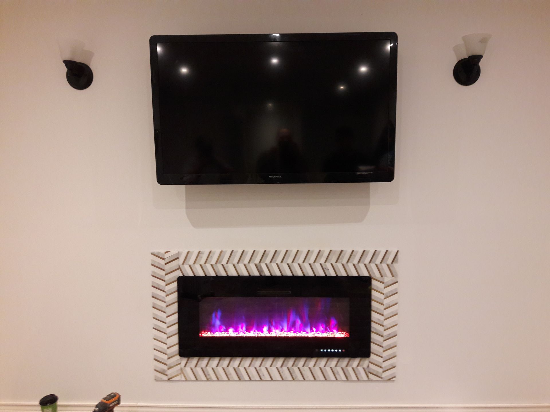 A wall-mounted TV sits above an electric fireplace surrounded by a white herringbone tile border, flanked by two sconces.