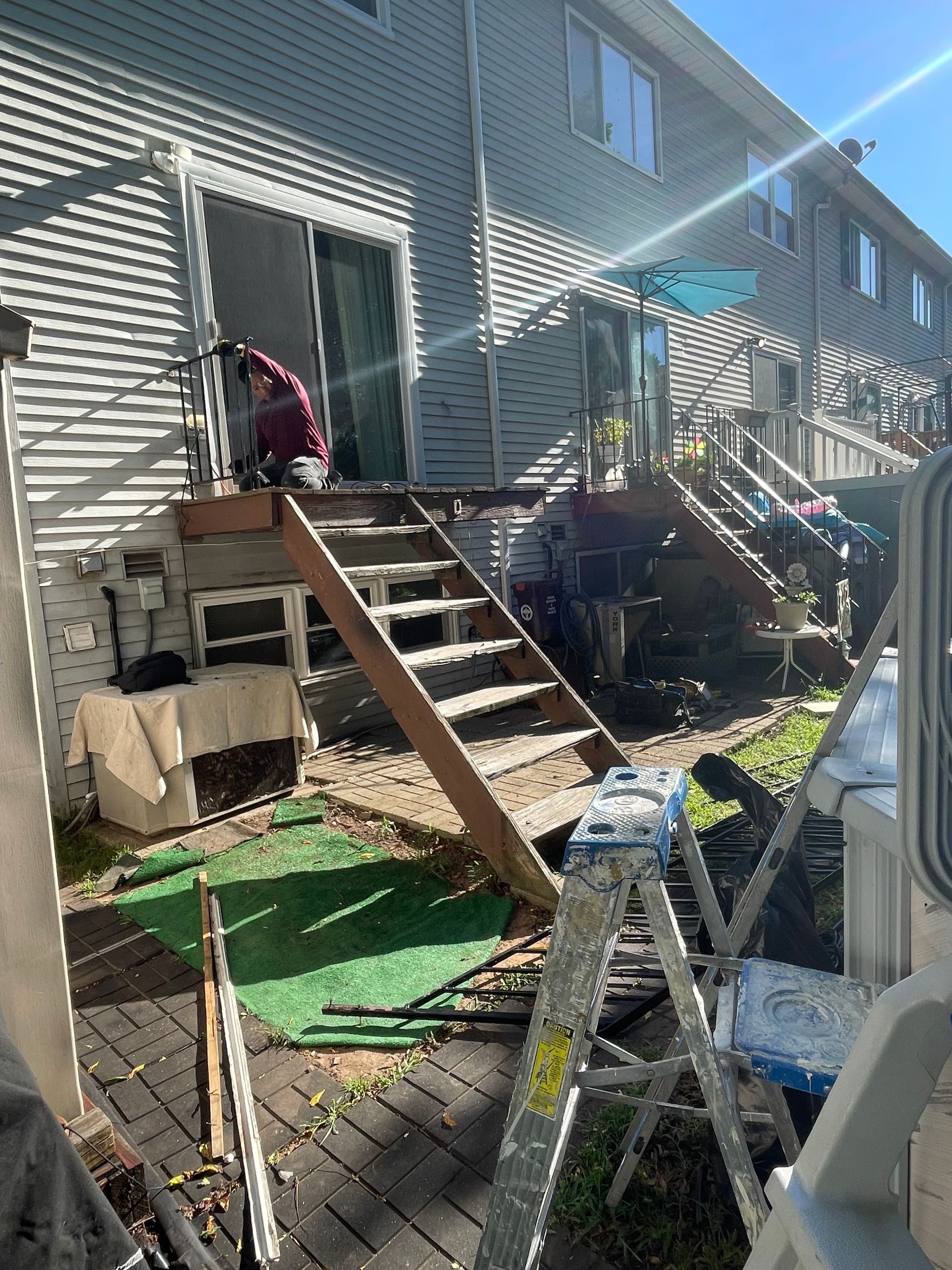 A person in a red jacket works on a sliding glass door at the top of a wooden deck staircase leading to a brick patio.