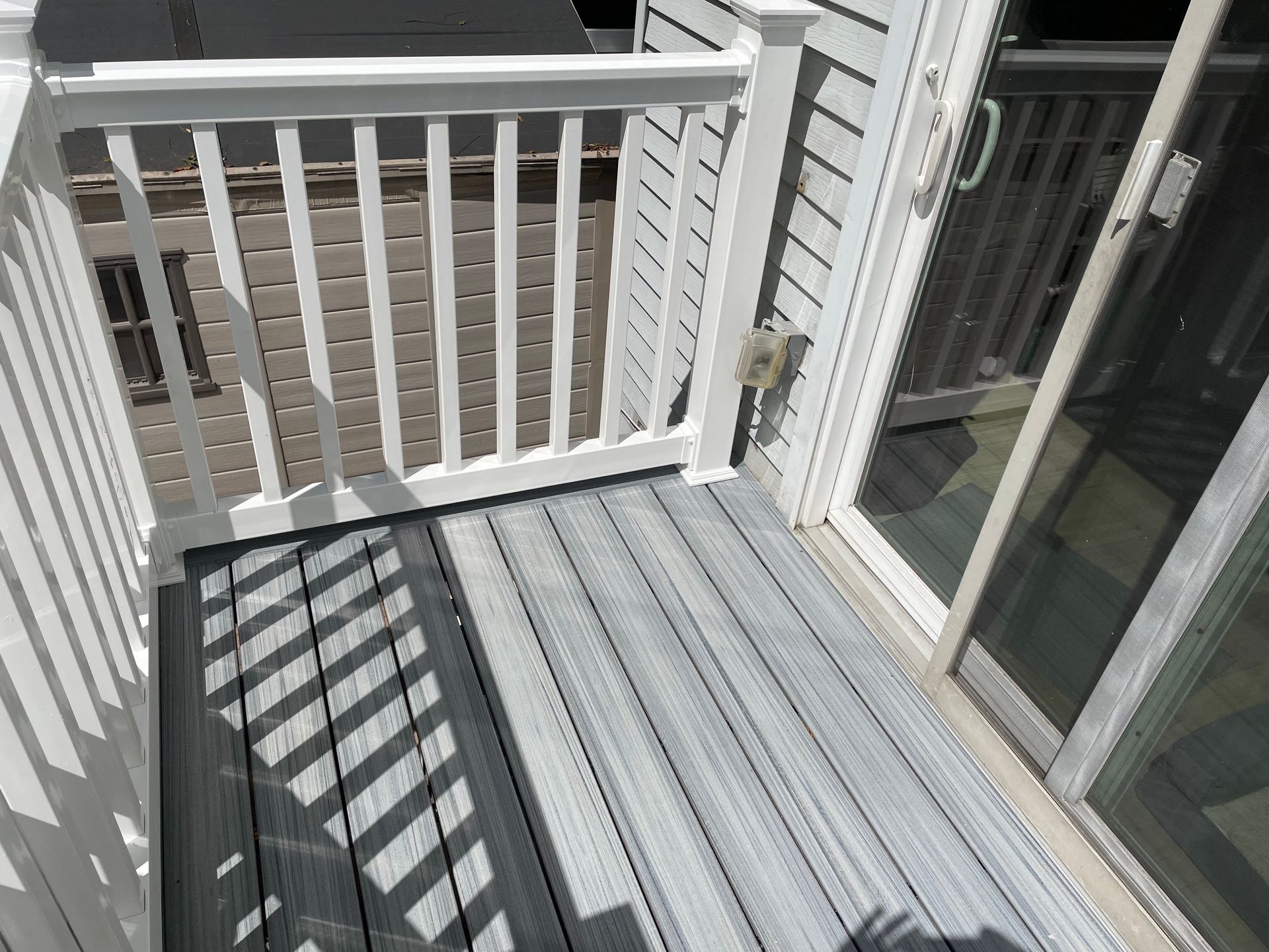A high-angle view of a grey wooden balcony deck with white railings, featuring a sliding glass door and side siding.