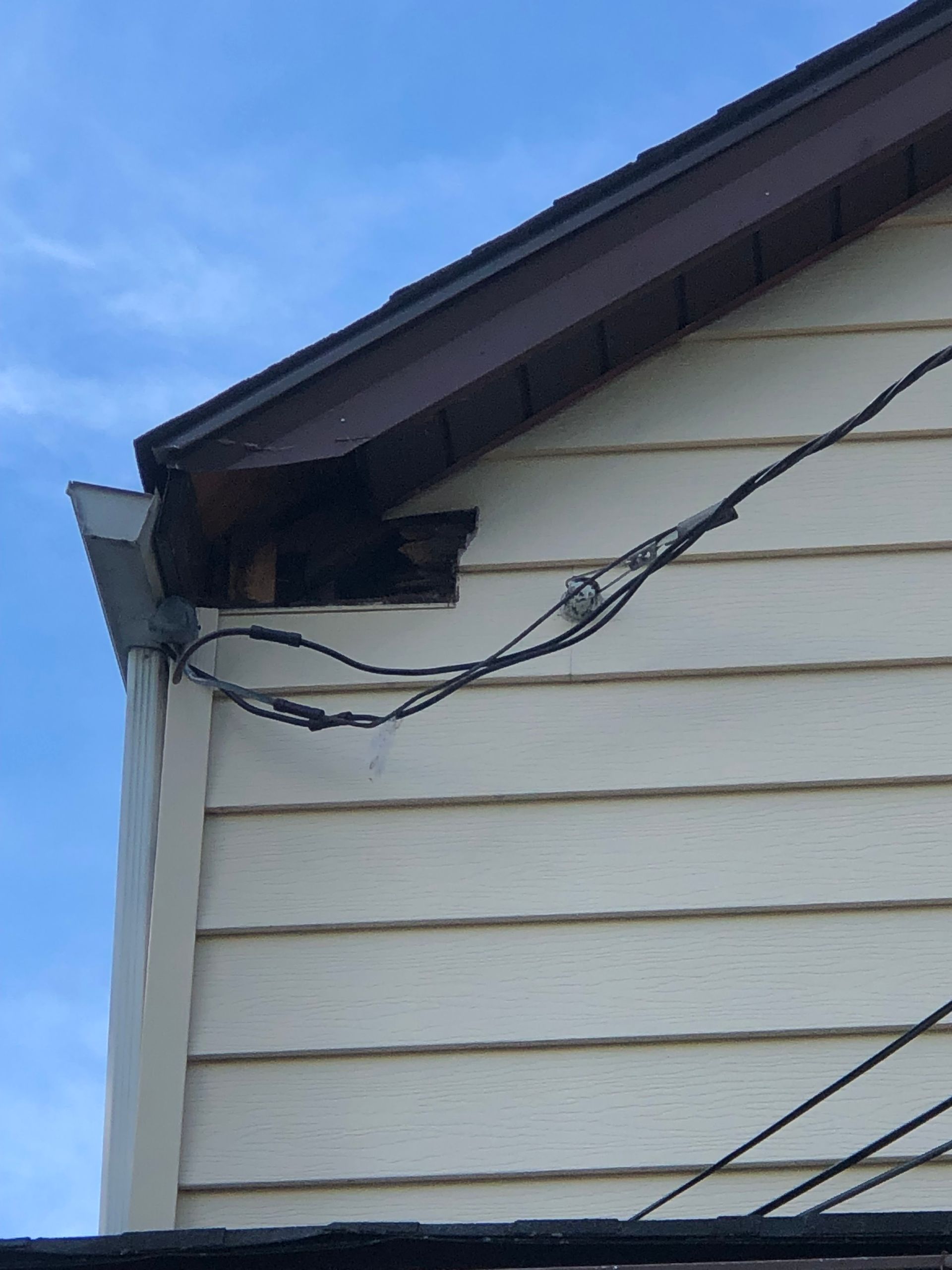 A gap in the exterior wall where a roof eave meets siding, showing dark, exposed wood and hanging utility wires.