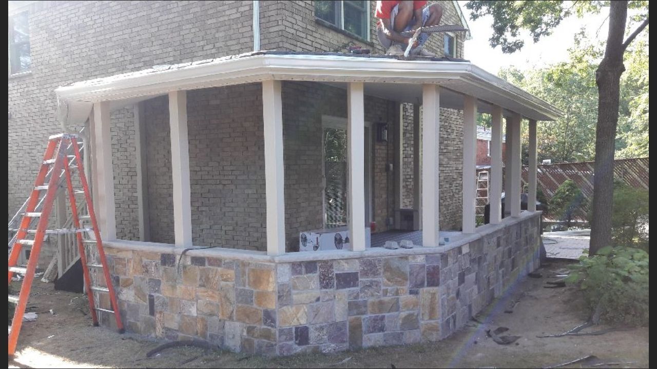 A worker on a roof installs a new covered patio with stone-veneer walls and white pillars attached to a residential house.