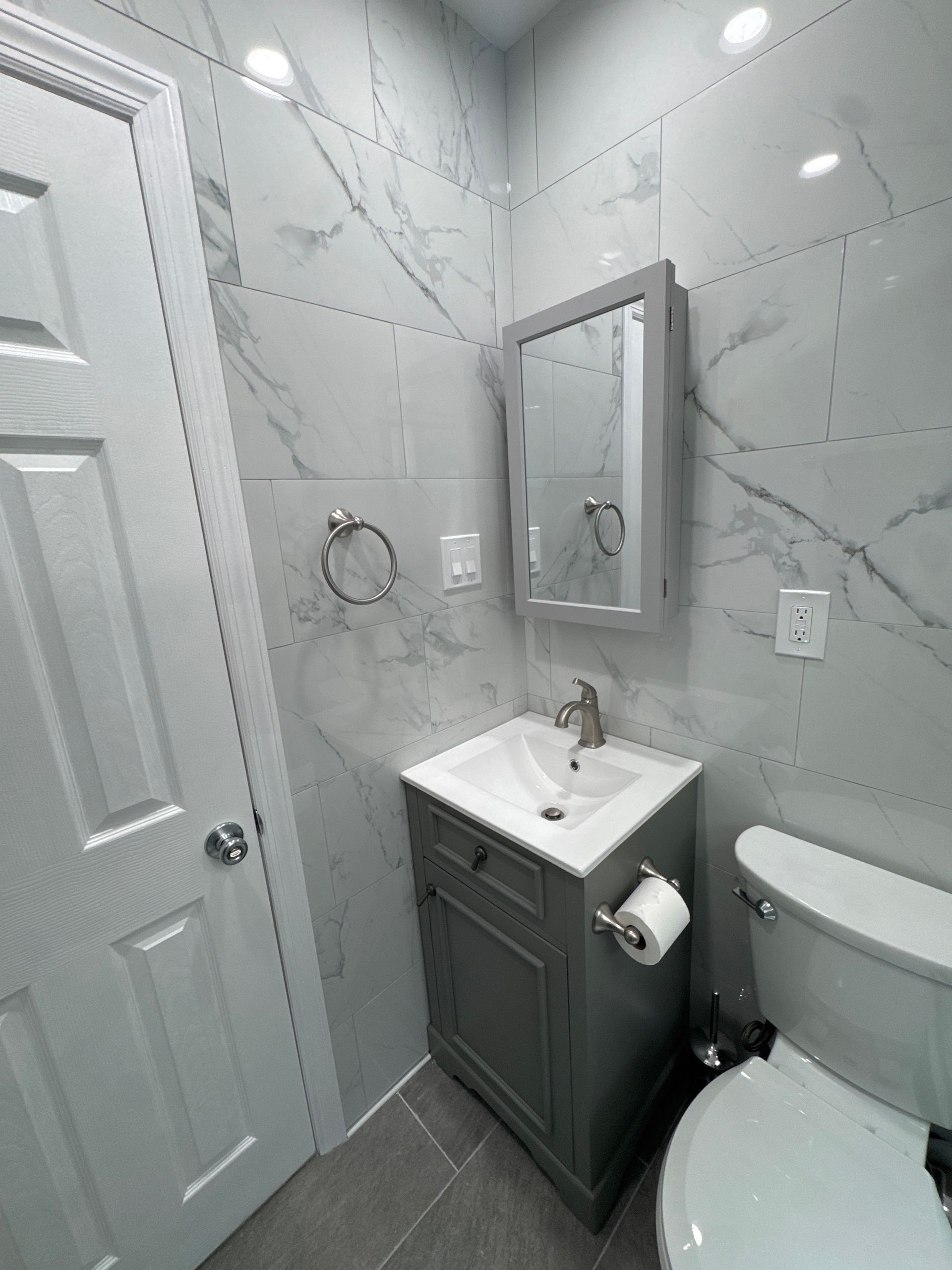 Small gray bathroom with tiled walls, vanity sink, mirror, towel ring, and toilet