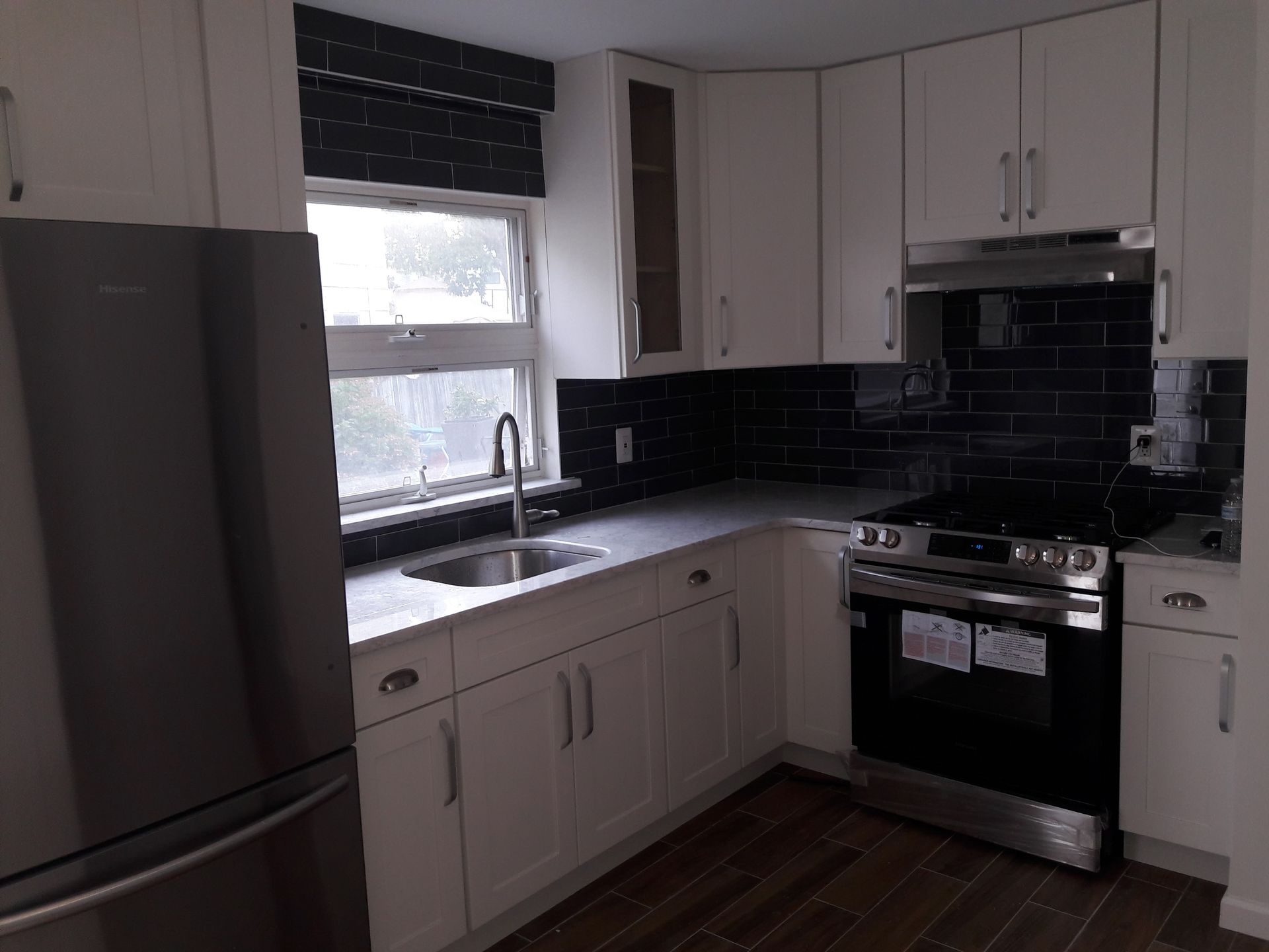A modern kitchen with white cabinets, dark subway tile backsplashes, a stainless steel refrigerator, and wood flooring.