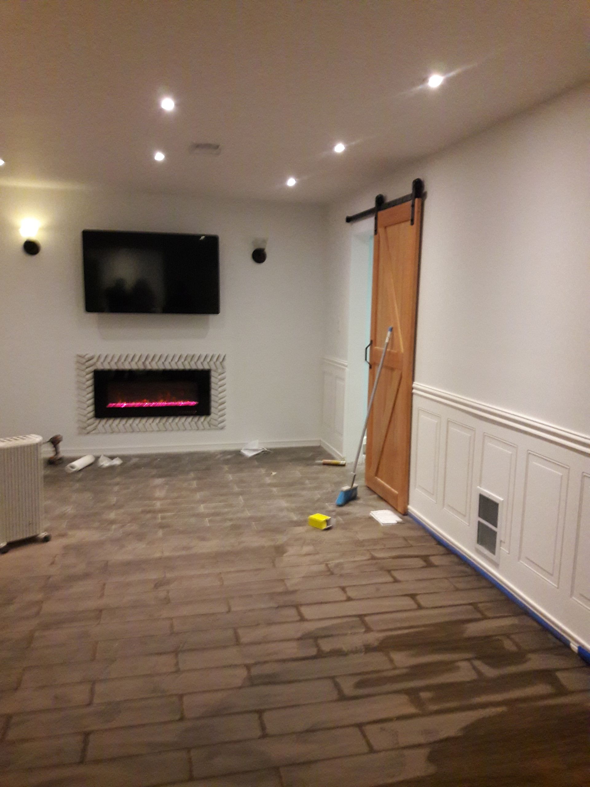 A living room with a fireplace, wall-mounted TV, wood-look tiled floor, sliding wooden door, and white wainscoting.