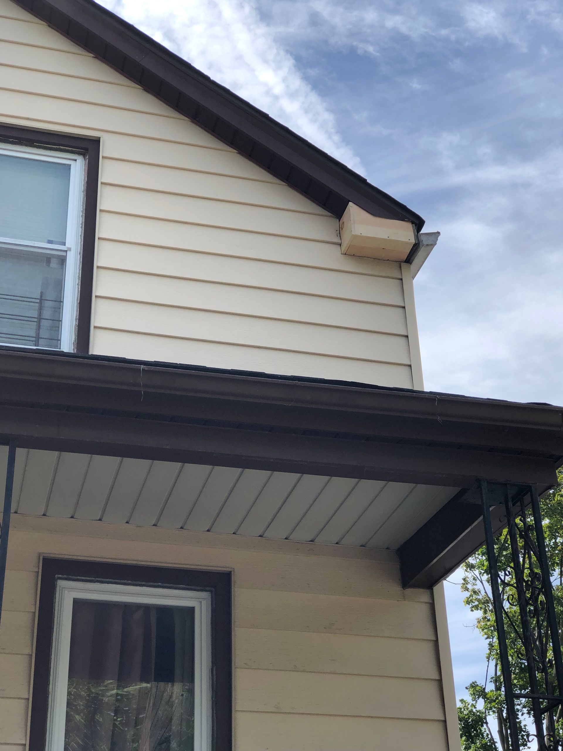 A two-story tan house exterior featuring a beige rectangular block attached to the roofline under the eaves.
