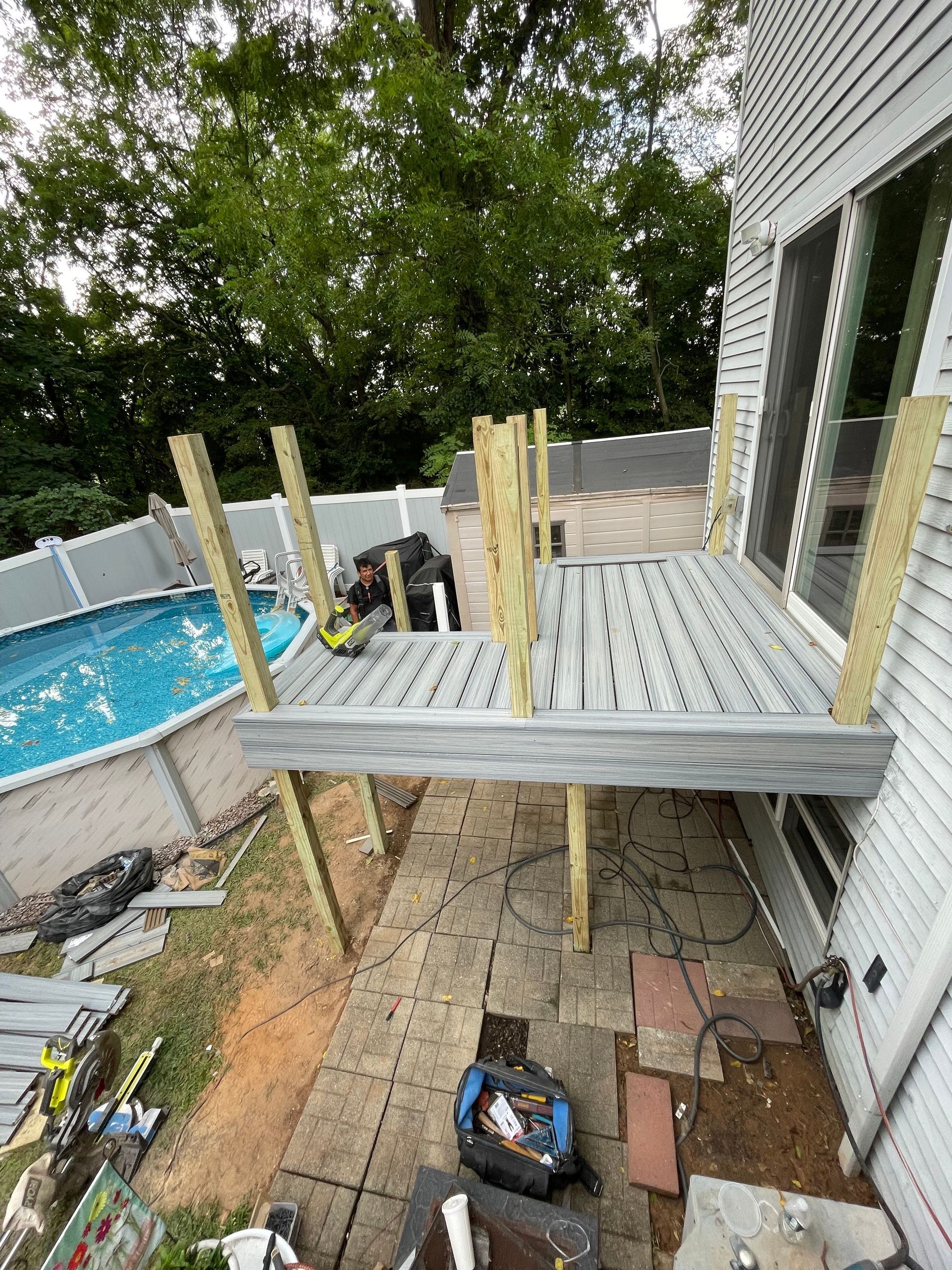 A small, unfinished wooden deck under construction next to a house, with vertical posts installed and a swimming pool.