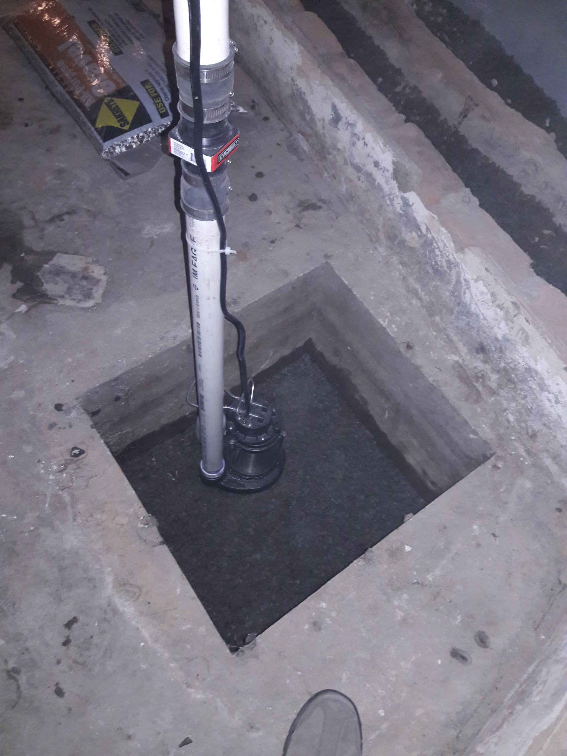 A sump pump with a vertical white PVC discharge pipe stands inside a square concrete basin in a basement floor.