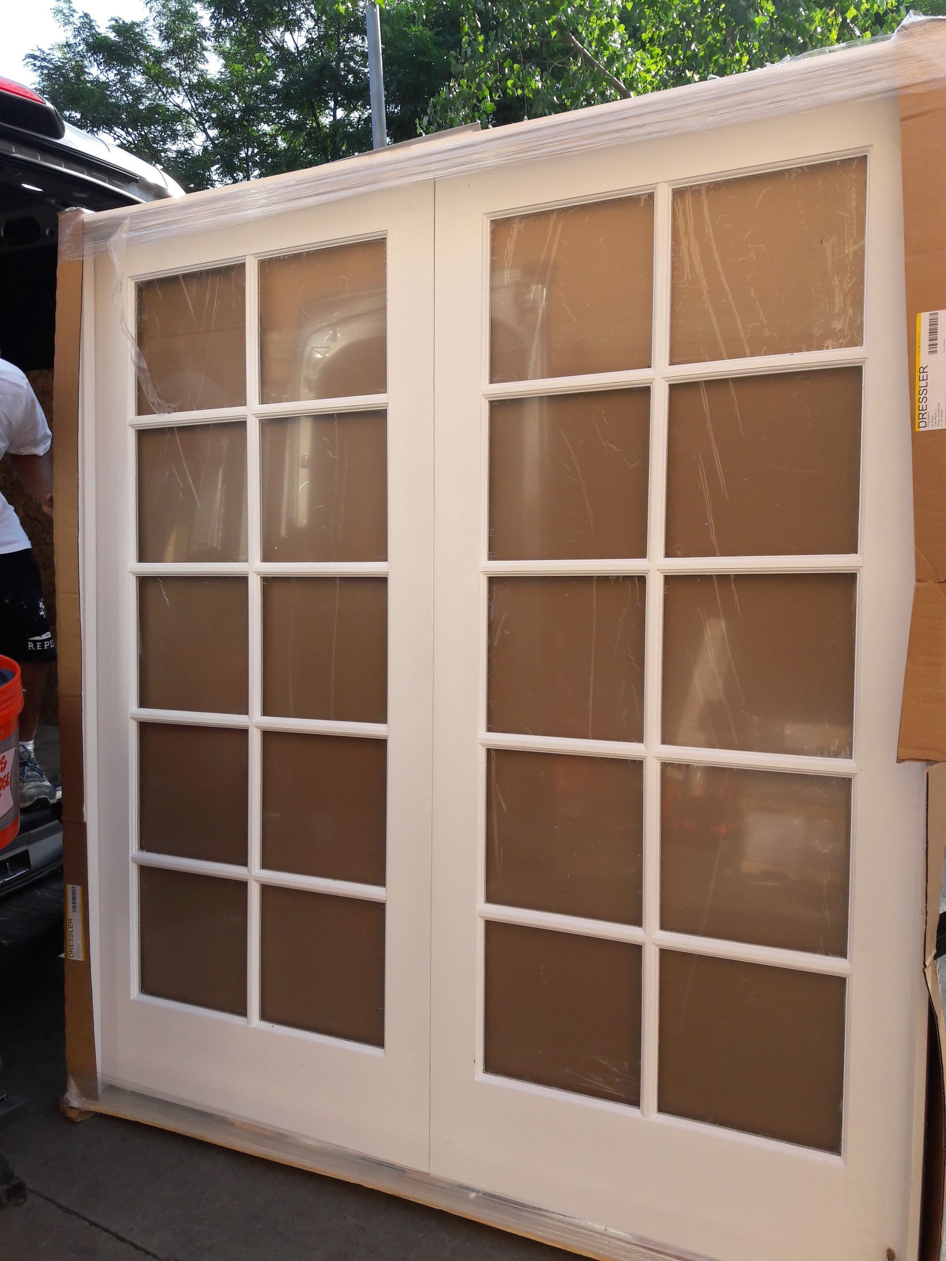 A set of white-framed French doors with ten glass panes per door, standing outdoors in cardboard packaging.
