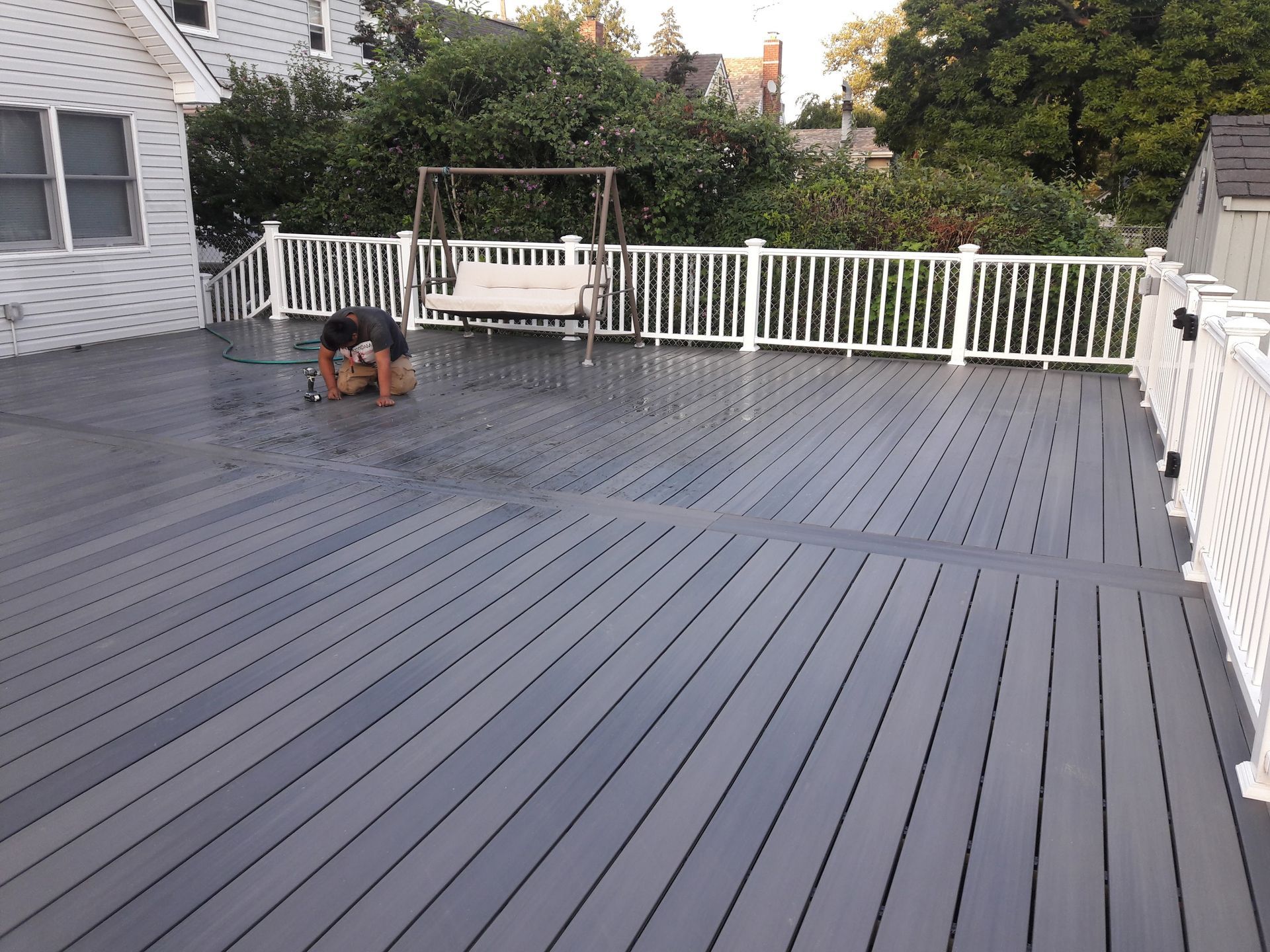 A person cleans a large, grey composite deck outdoors next to a house with a white railing and a swing.