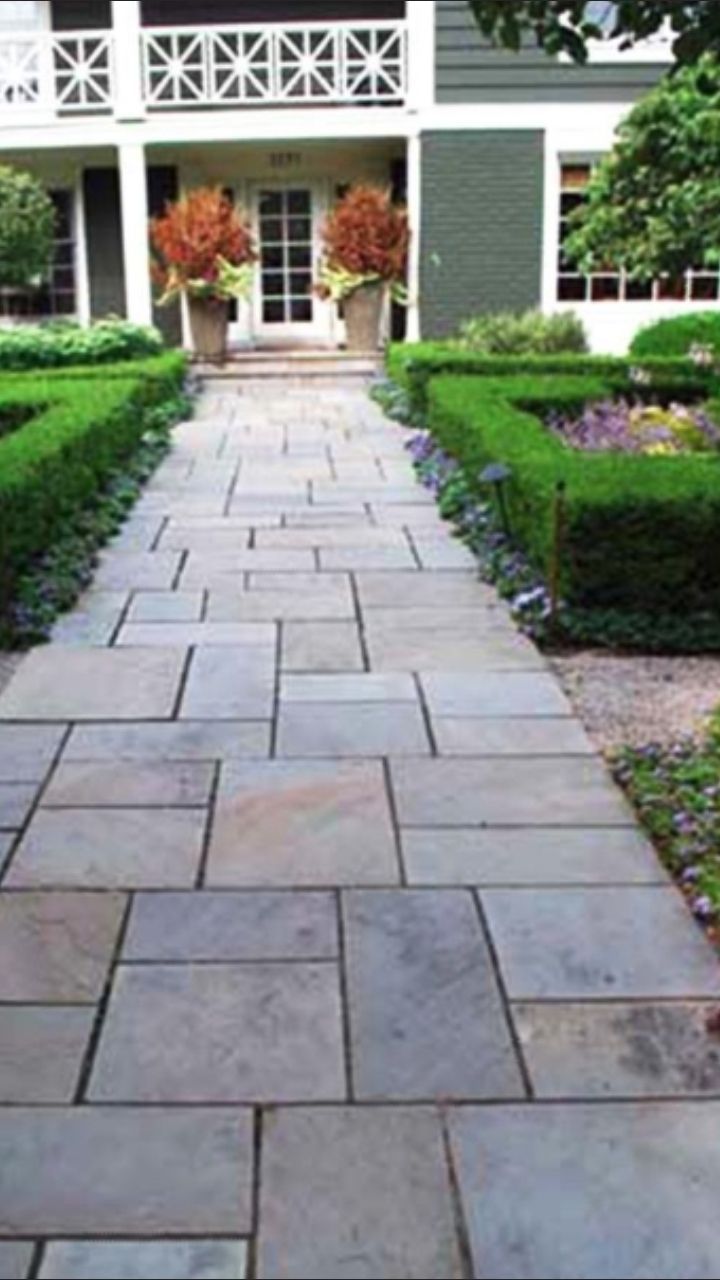 A stone pathway leads to the front entrance of a house, flanked by manicured green hedges and potted autumn-toned plants.