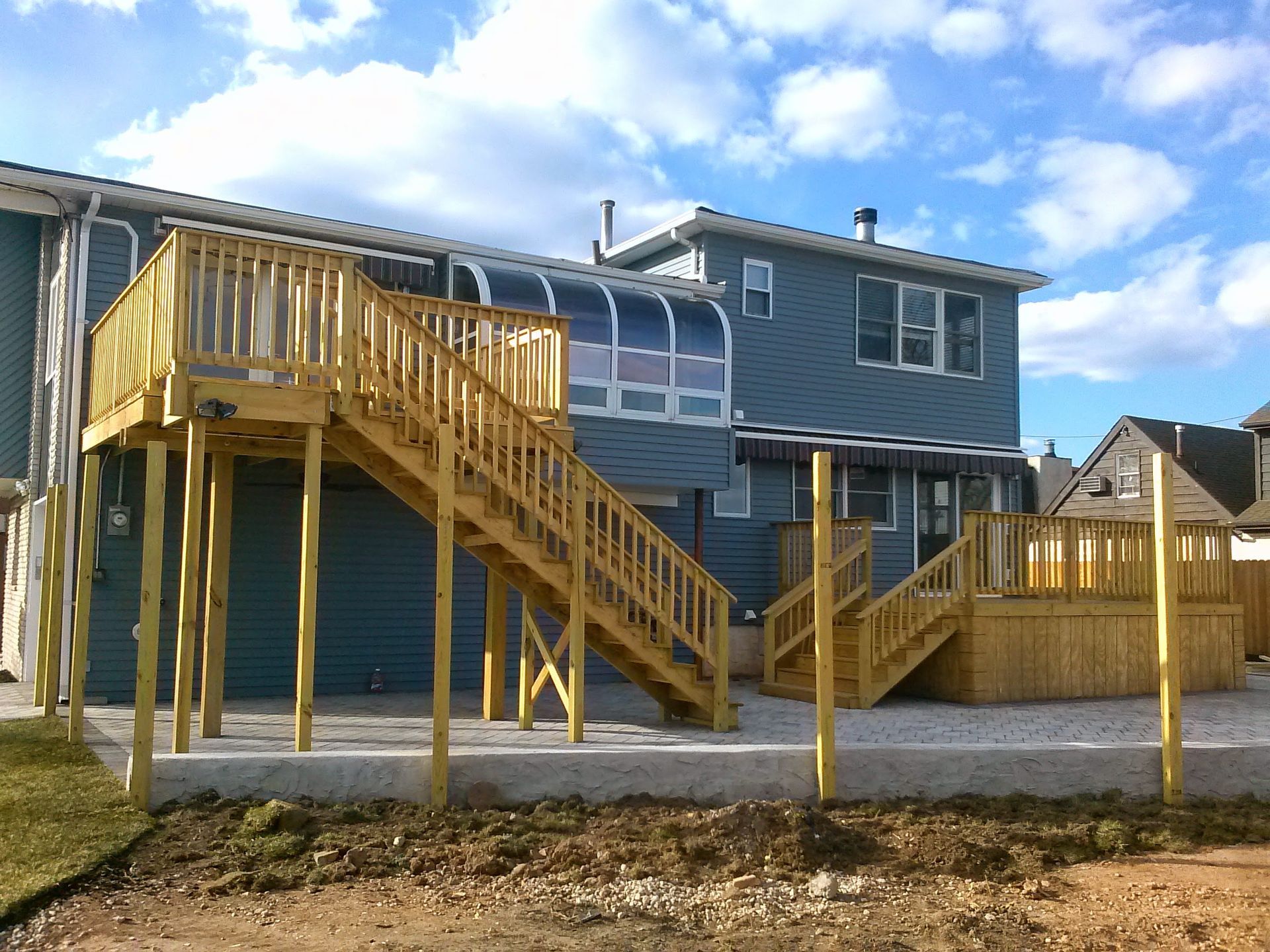 A two-story blue house with a newly constructed wooden deck and staircase system over a paved patio area.