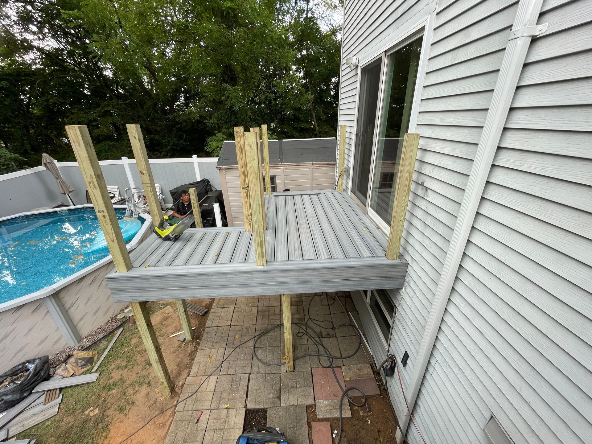 A wooden deck under construction is attached to a house next to an above-ground swimming pool in a backyard.