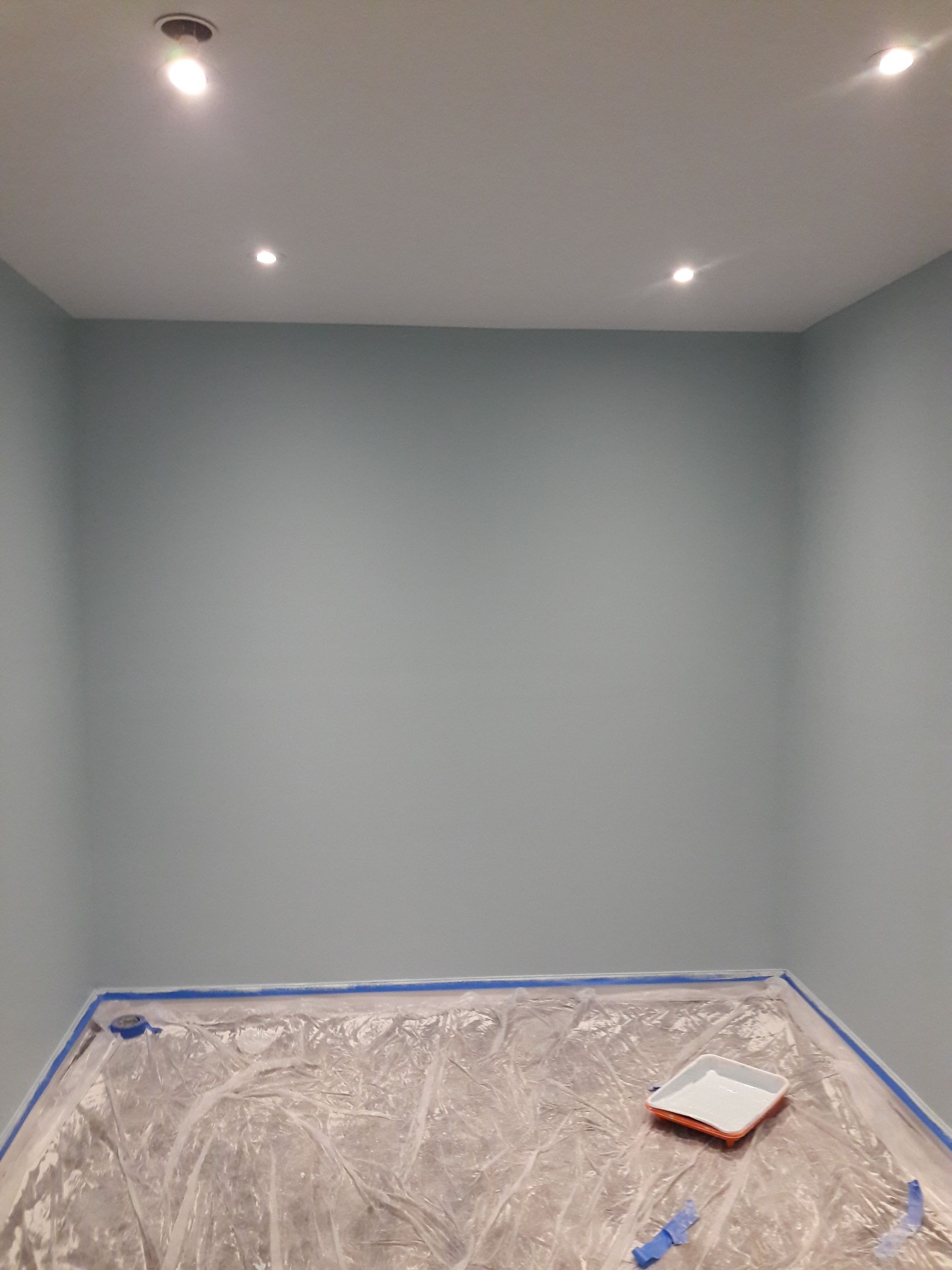 A freshly painted light blue room with four recessed ceiling lights and a paint tray on a plastic-covered floor.