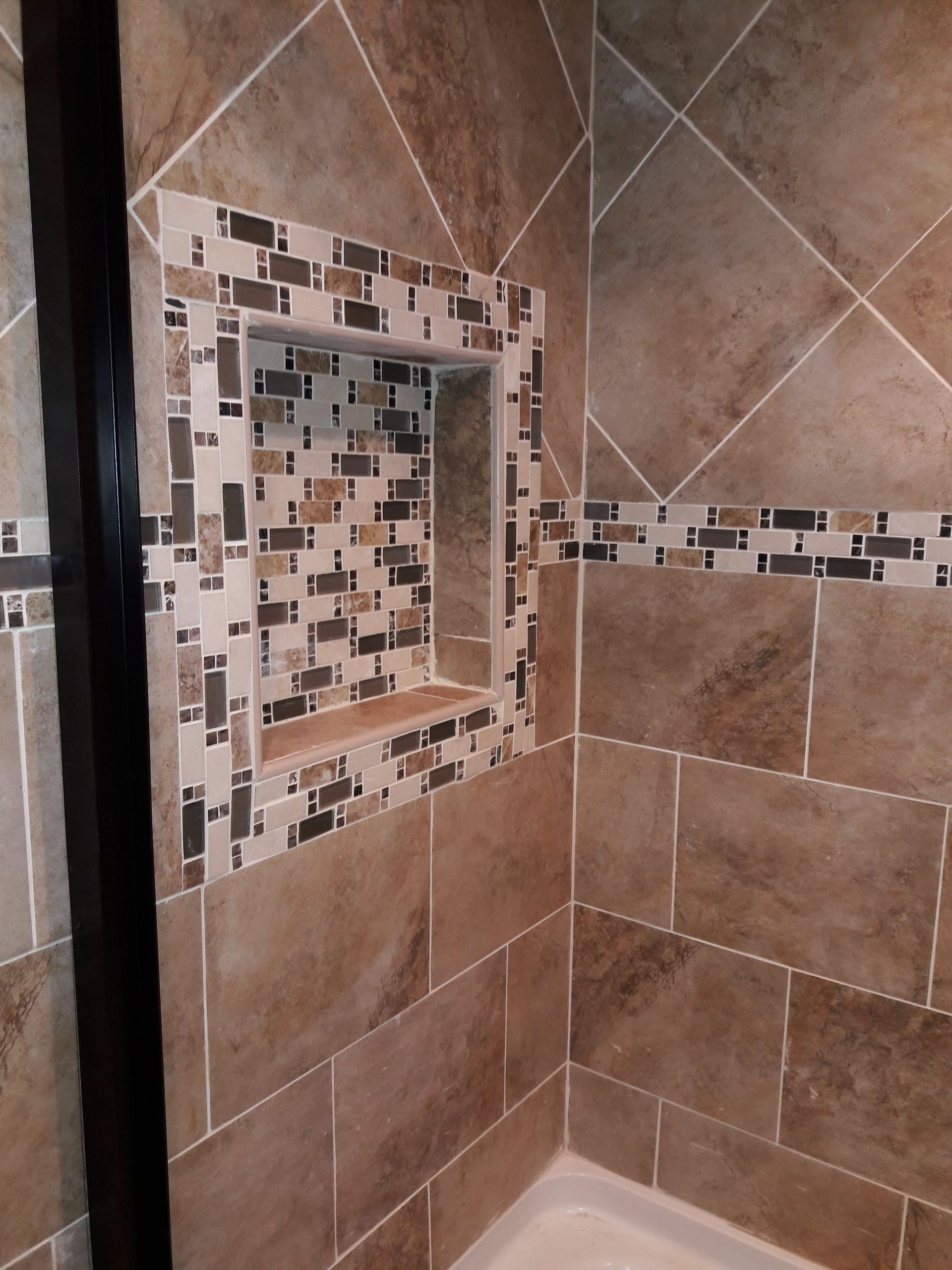 A shower wall tiled in brown stone with a built-in mosaic-tiled niche and a horizontal accent strip.