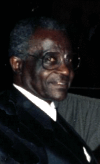 Smiling man in a dark suit and glasses, seated against a dark background.