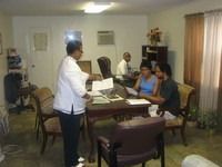 Two people seated at a table while a woman stands and speaks in a small meeting room.