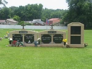 Cemetery memorial with headstones and flowers on a grassy lawn, with houses and trees in the background