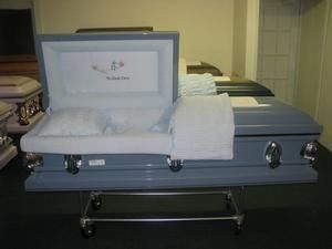 Open casket with a white satin interior and folded white blanket on a metal stand.