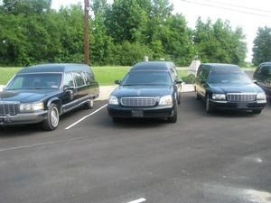 Three black hearses parked side by side in a lot, with trees in the background.