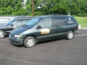 Dark green minivan with company logo parked on asphalt beside another car, trees in the background
