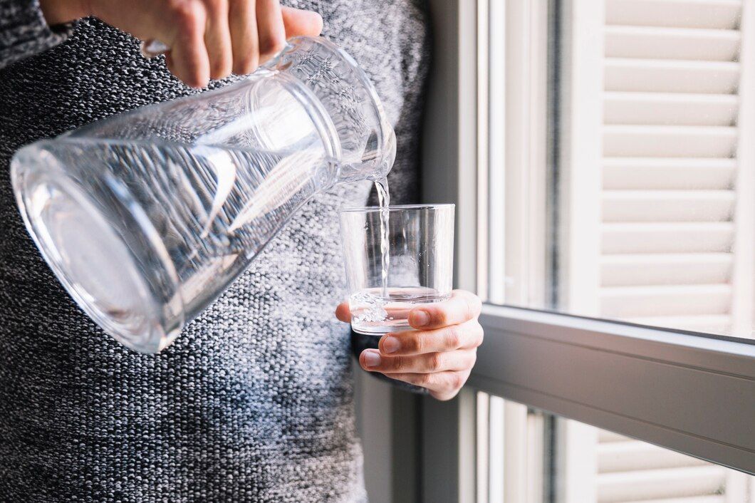 Person pouring water from a pitcher into a glass by a window.