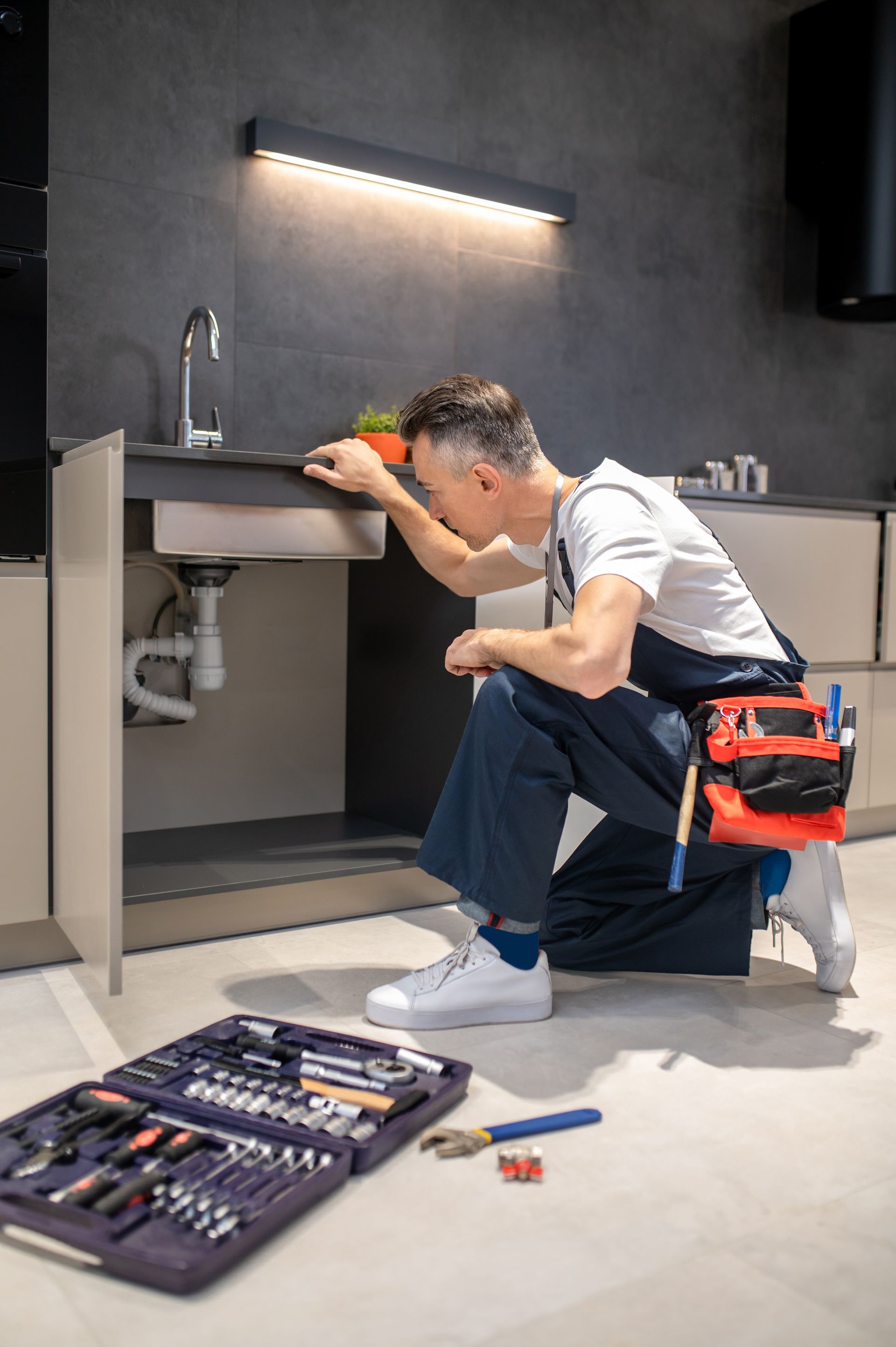 Plumber crouched under kitchen sink, examining pipes. Tool kit and tools on floor.