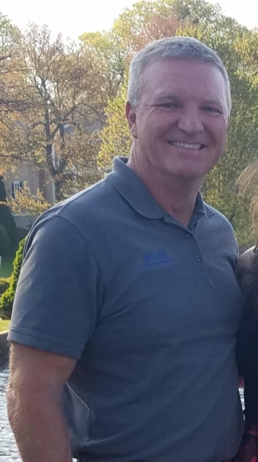 Man in gray polo shirt smiles outdoors with trees in the background.