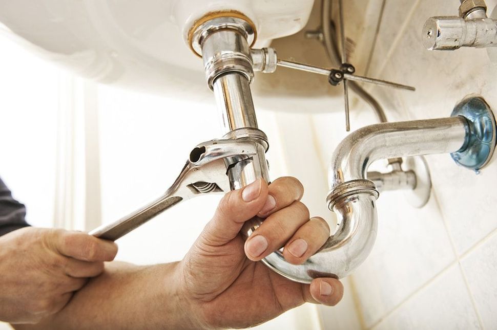Plumber using a wrench to tighten a pipe under a white sink in a bathroom.