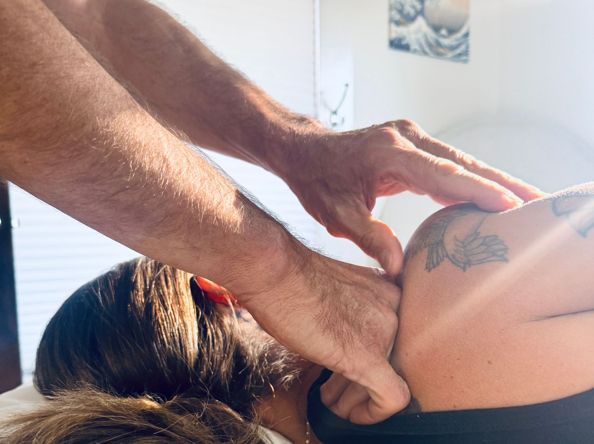 Hands massaging a person's shoulder and neck. Person lying down with dark hair. Bright light, indoor setting.