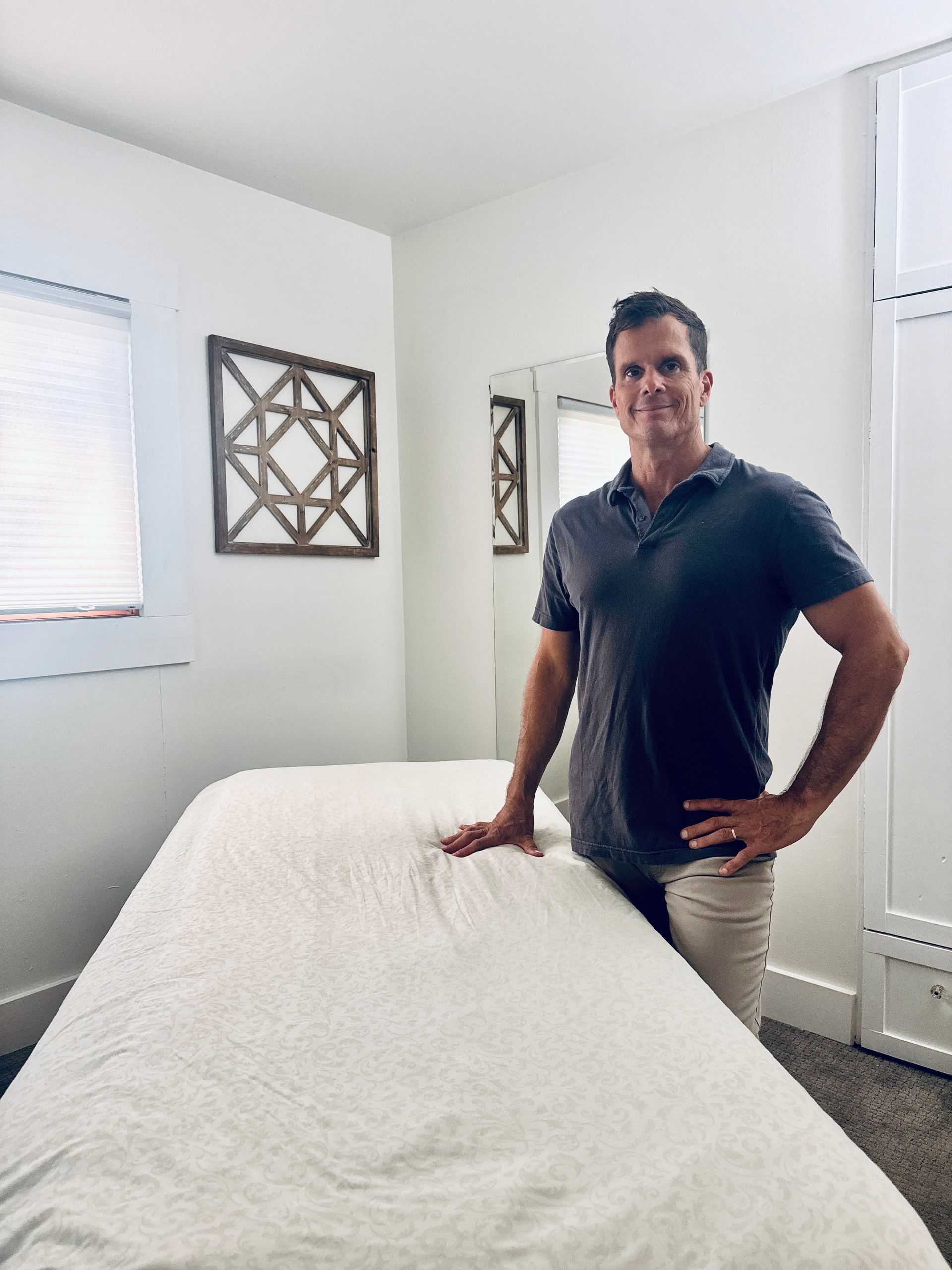 Man stands beside massage table in a light-filled room; hand on the table, smiling.