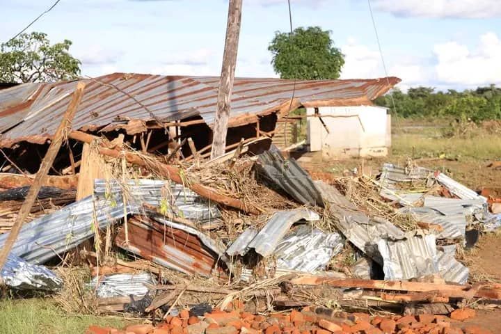 A collapsed building with twisted metal roofing, wooden debris, and scattered bricks in a rural, open-field setting.
