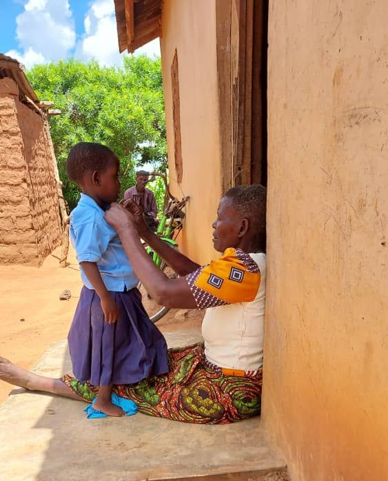 A person sits on a porch, adjusting the collar of a child's school uniform against the wall of a mud-brick home.