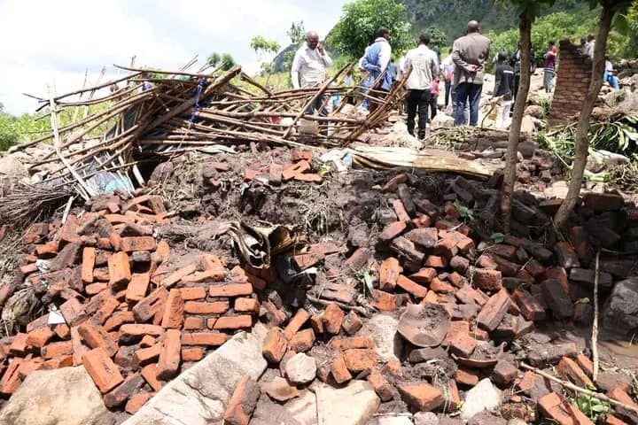 A group of people standing on a hillside overlooking the wreckage of a collapsed red brick building and scattered debris.