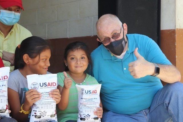 A man wearing a mask is giving a thumbs up next to two little girls holding usaid bags