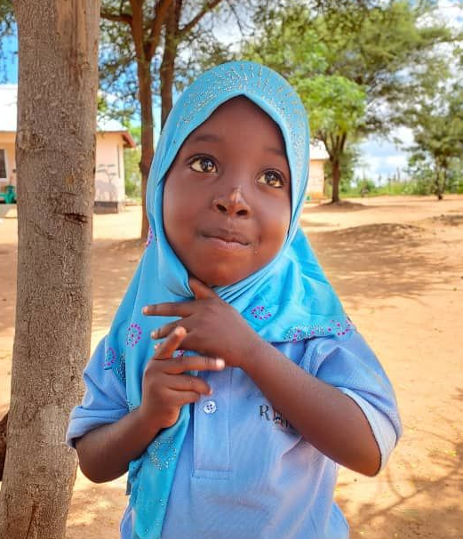 A child wearing a light blue hijab and shirt, smiling while touching their chin, standing outdoors in a sandy area.