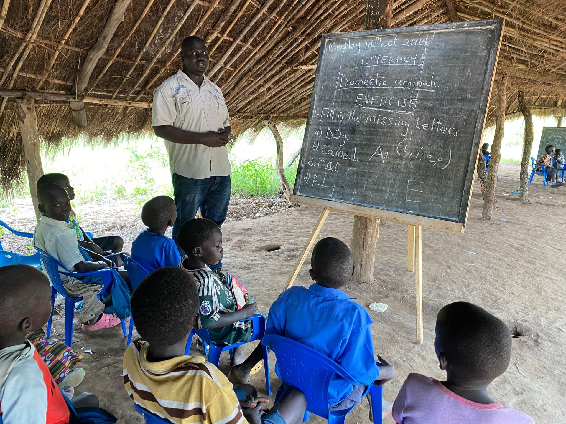 A group of children are sitting in chairs under a thatched roof while a man stands behind a blackboard.