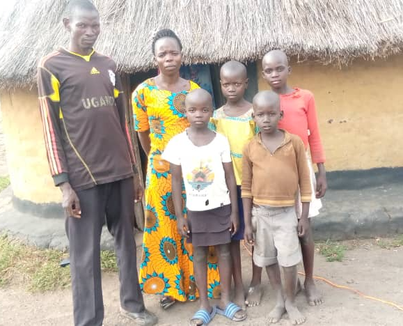 A group of people standing in front of a thatched hut with a man wearing an uganda shirt