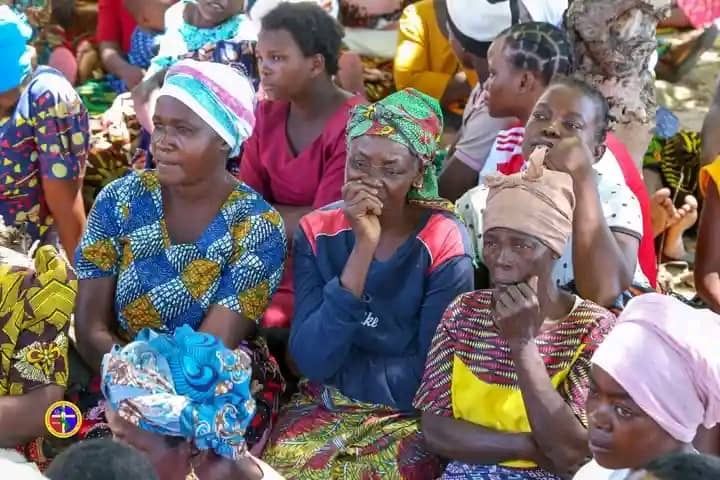 A group of people sitting outdoors, wearing colorful headscarves and patterned clothing, with serious expressions.