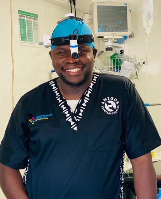 A smiling healthcare professional in dark scrubs and a headlamp stands in a clinical setting with medical equipment.