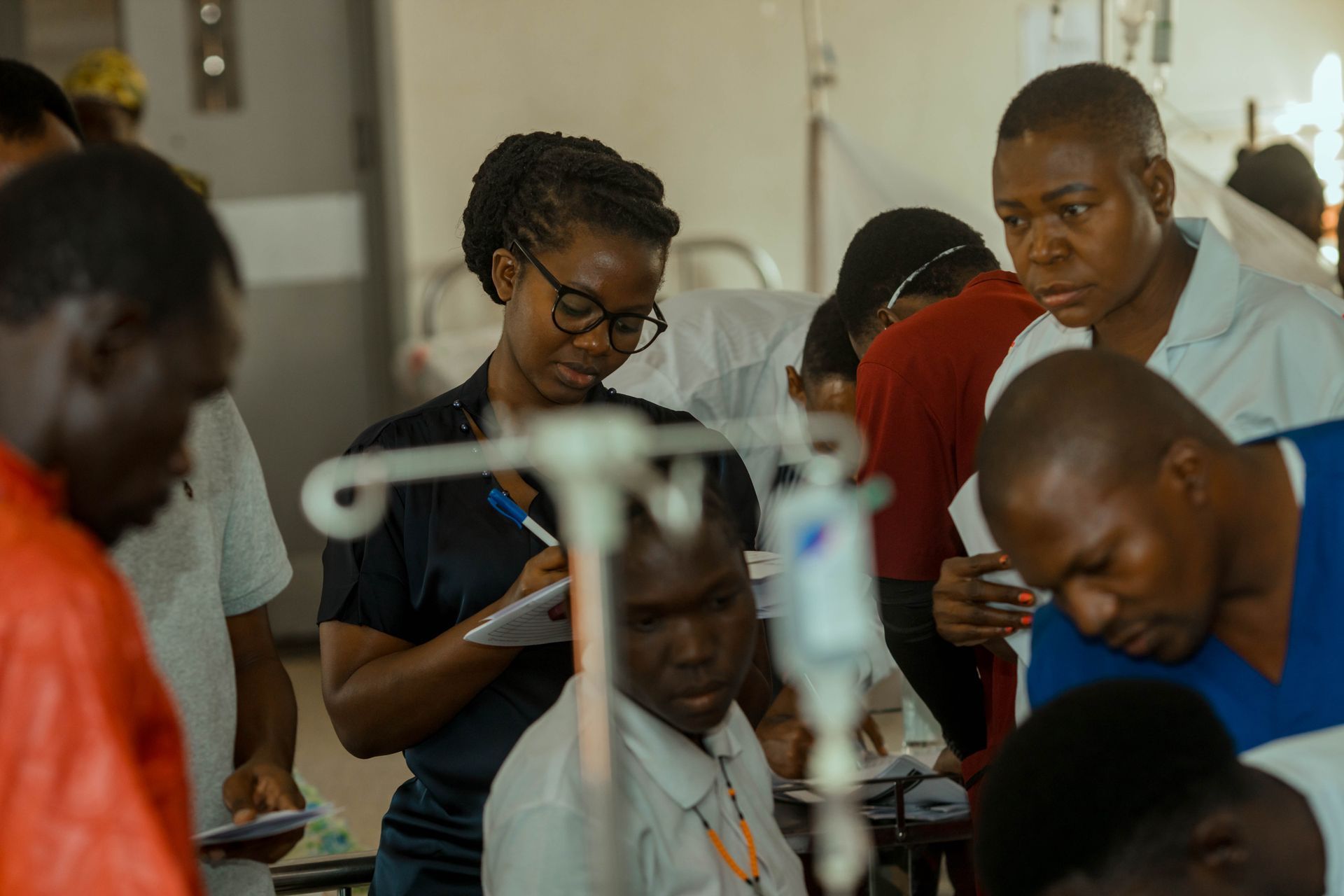 Medical staff tending to patients in a busy hospital ward; some taking notes, others looking on.