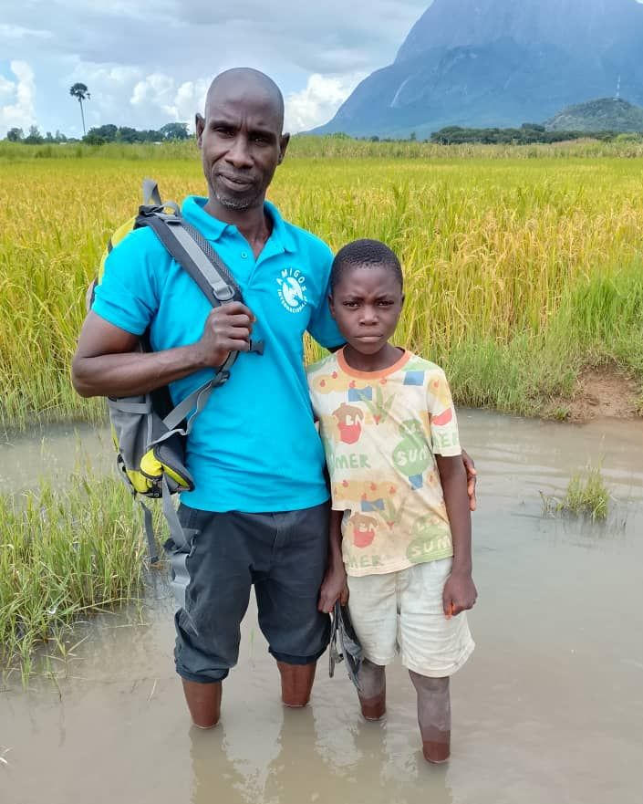 Two people stand in shallow water before a field of golden crops and a mountain, wearing casual clothes and a backpack.
