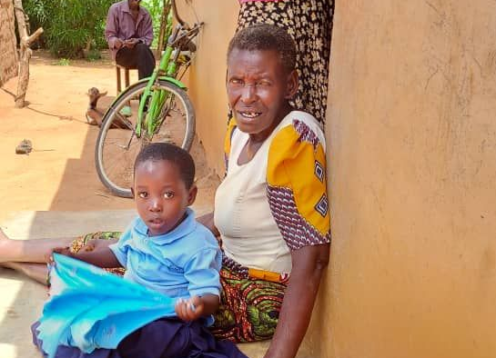 An older adult and a young child sit together on a dirt ground near a bicycle and a tan wall.