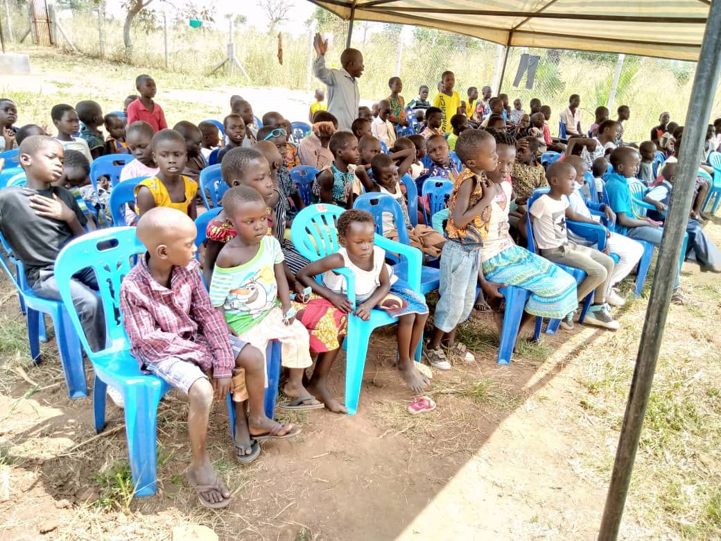 A group of children are sitting in blue chairs under a tent.