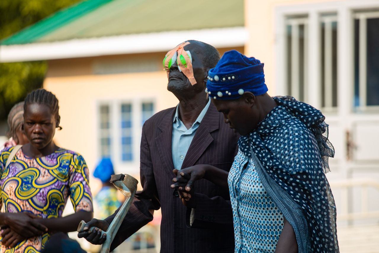 A group of people outside. A man with eye bandages leans on a crutch. A woman in blue beside him.
