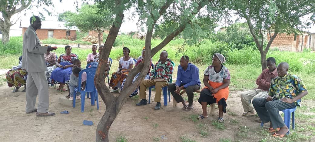 A man is standing in front of a group of people sitting under a tree.